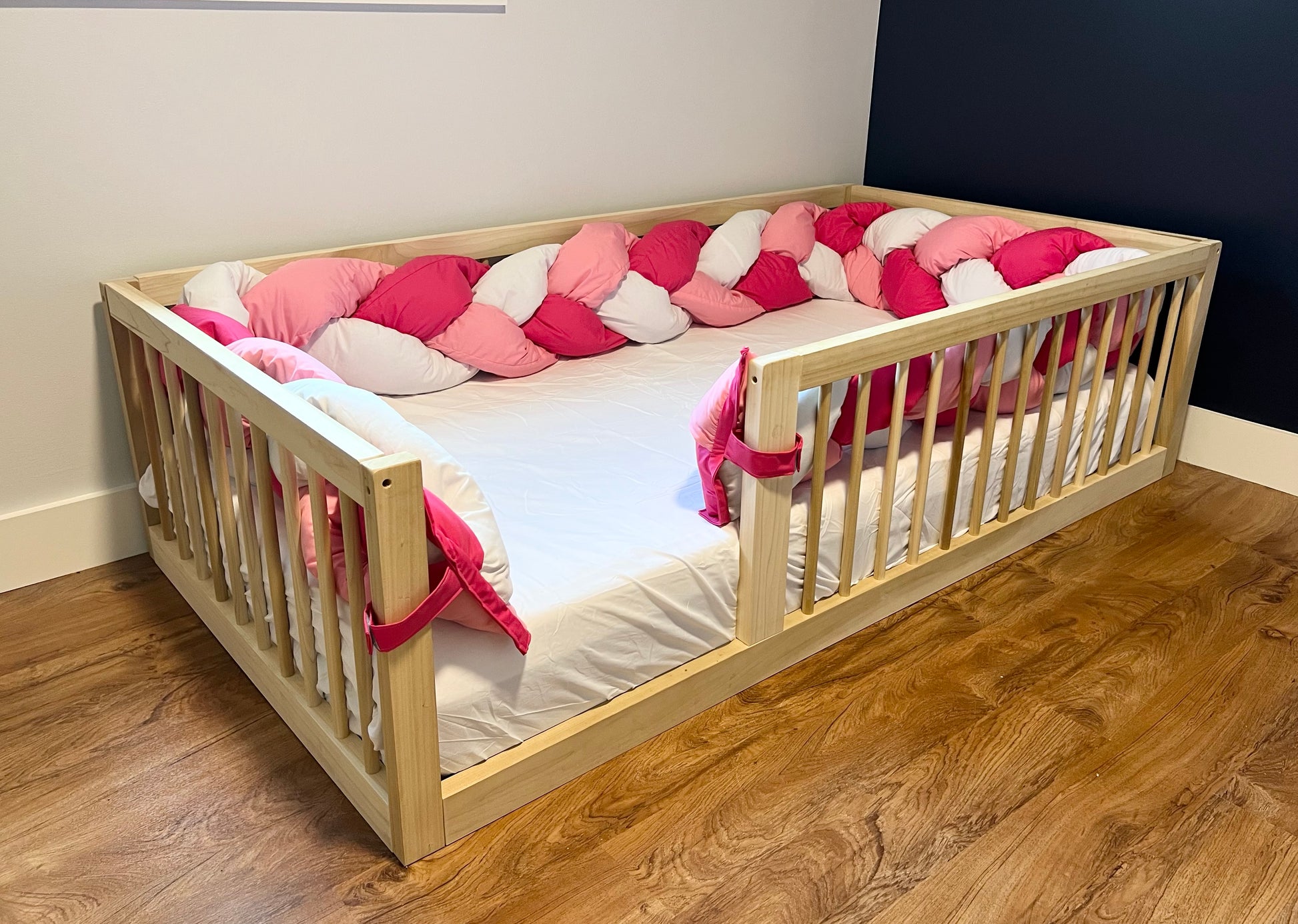 Wooden non-toxic montessori floor bed with pink and white braided bumper pillow.